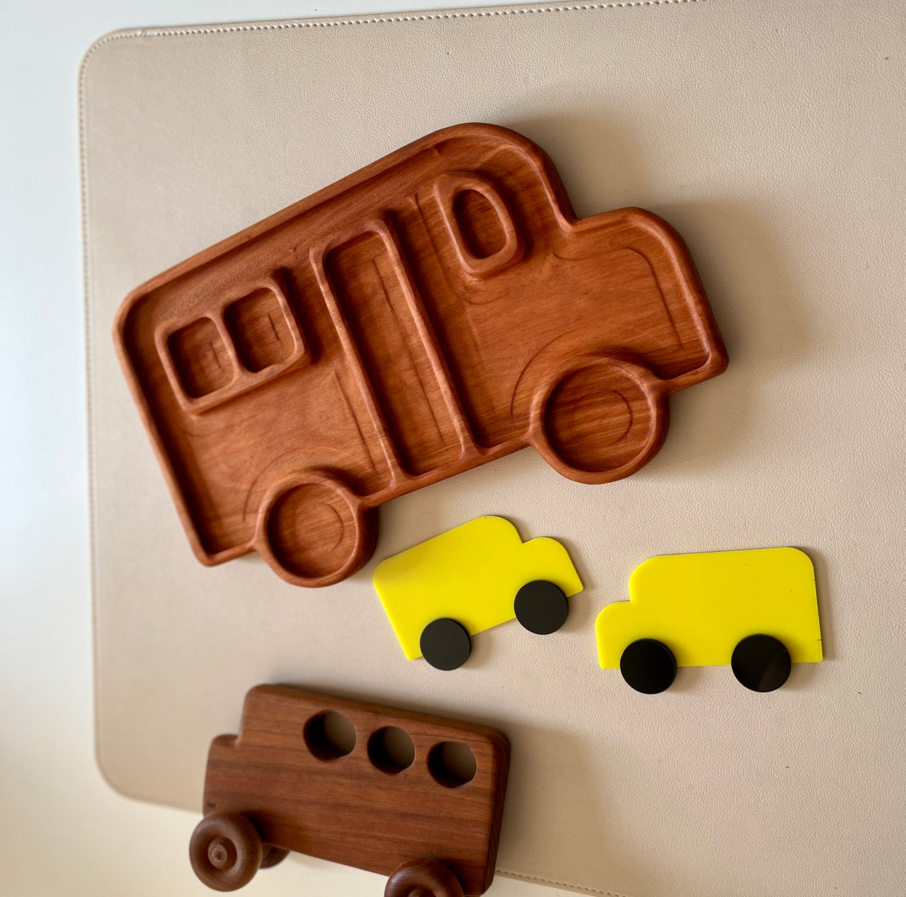 Wooden tray shaped like a camper van with two small yellow toy vans and a dark wooden bus toy on a light surface