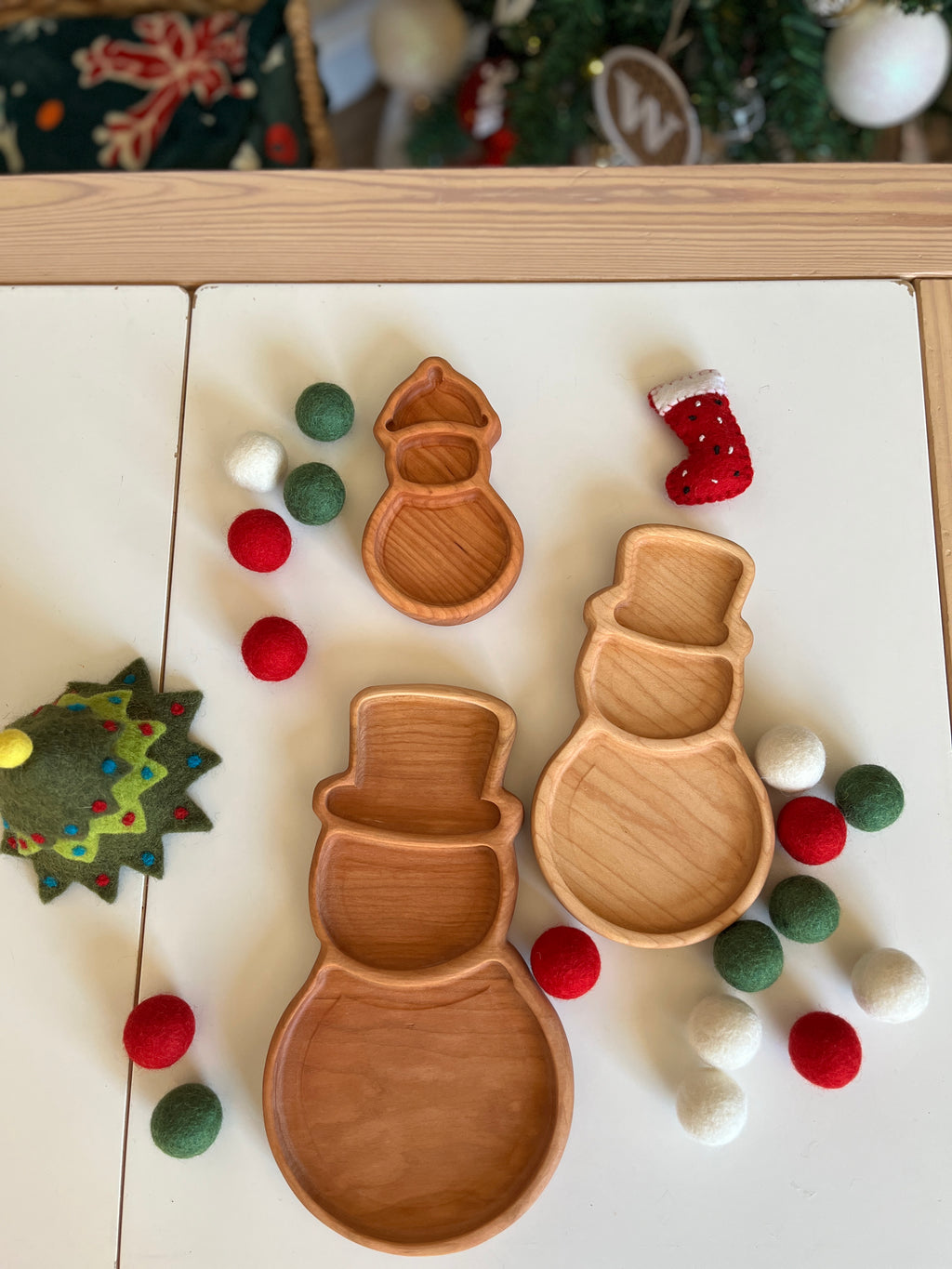 Three wooden snowman-shaped trays surrounded by red, white, and green felt balls and Christmas-themed felt decorations on table