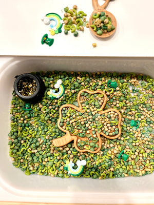 Sensory bin filled with green and beige pasta, wooden shamrock scoop, gold confetti, and rainbow and leprechaun charms