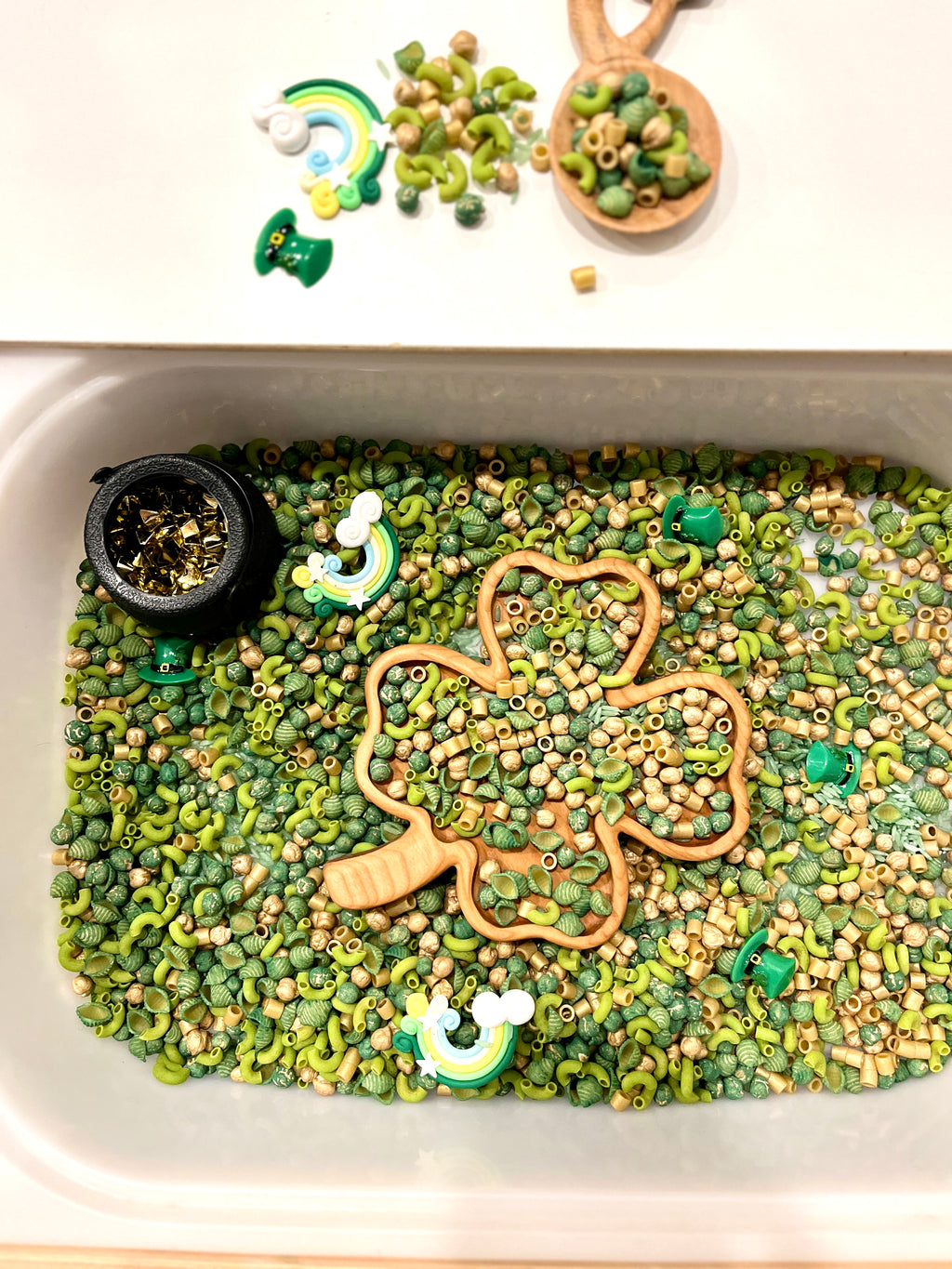 Sensory bin filled with green and beige pasta, wooden shamrock scoop, gold confetti, and rainbow and leprechaun charms