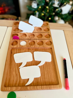 Wooden game board with blank white speech bubble cutouts, colored game pieces, and a pink marker on white surface
