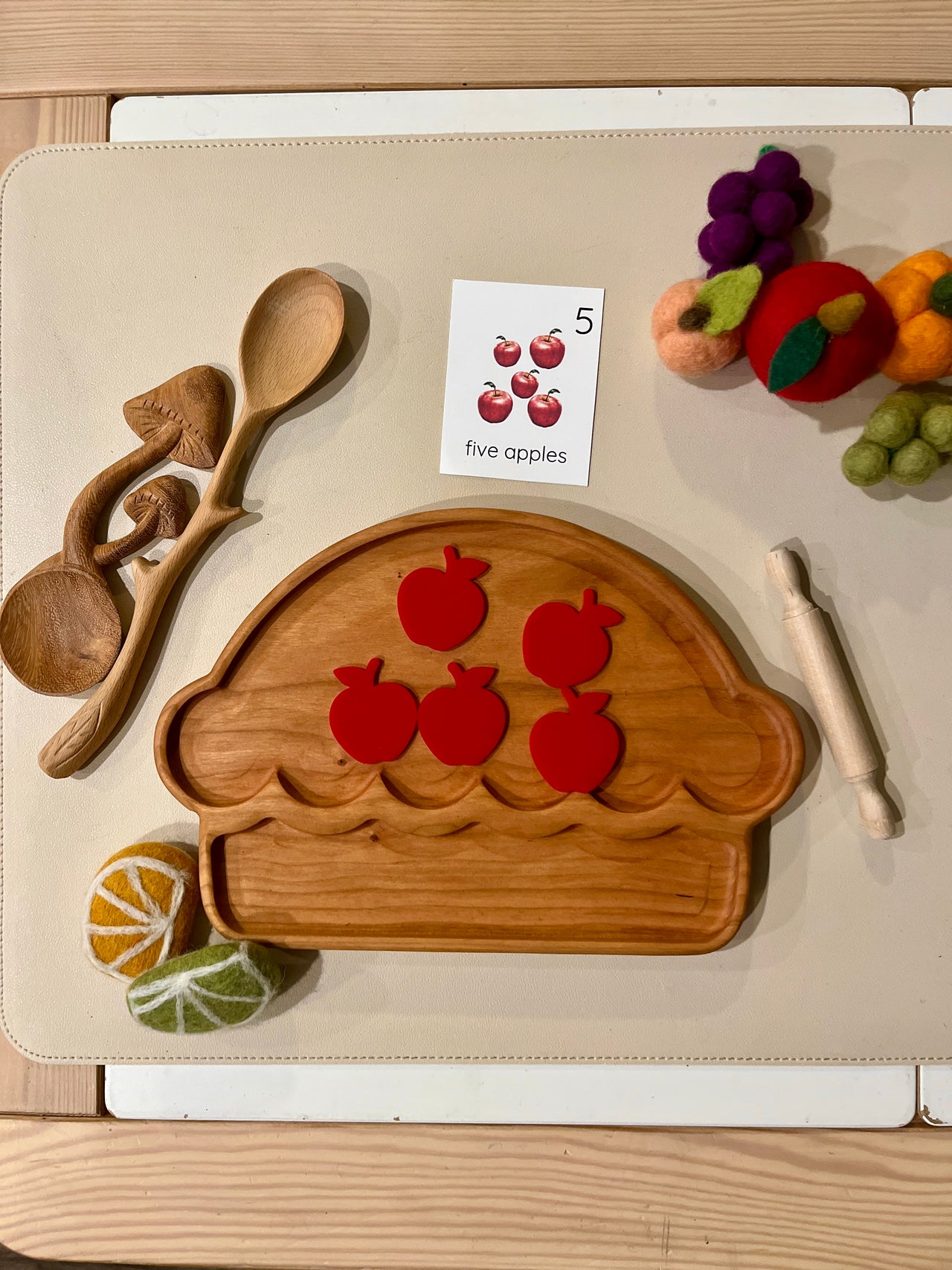 Wooden muffin-shaped tray with five red apple cutouts, wooden utensils, rolling pin, educational card, and felt fruit pieces nearby