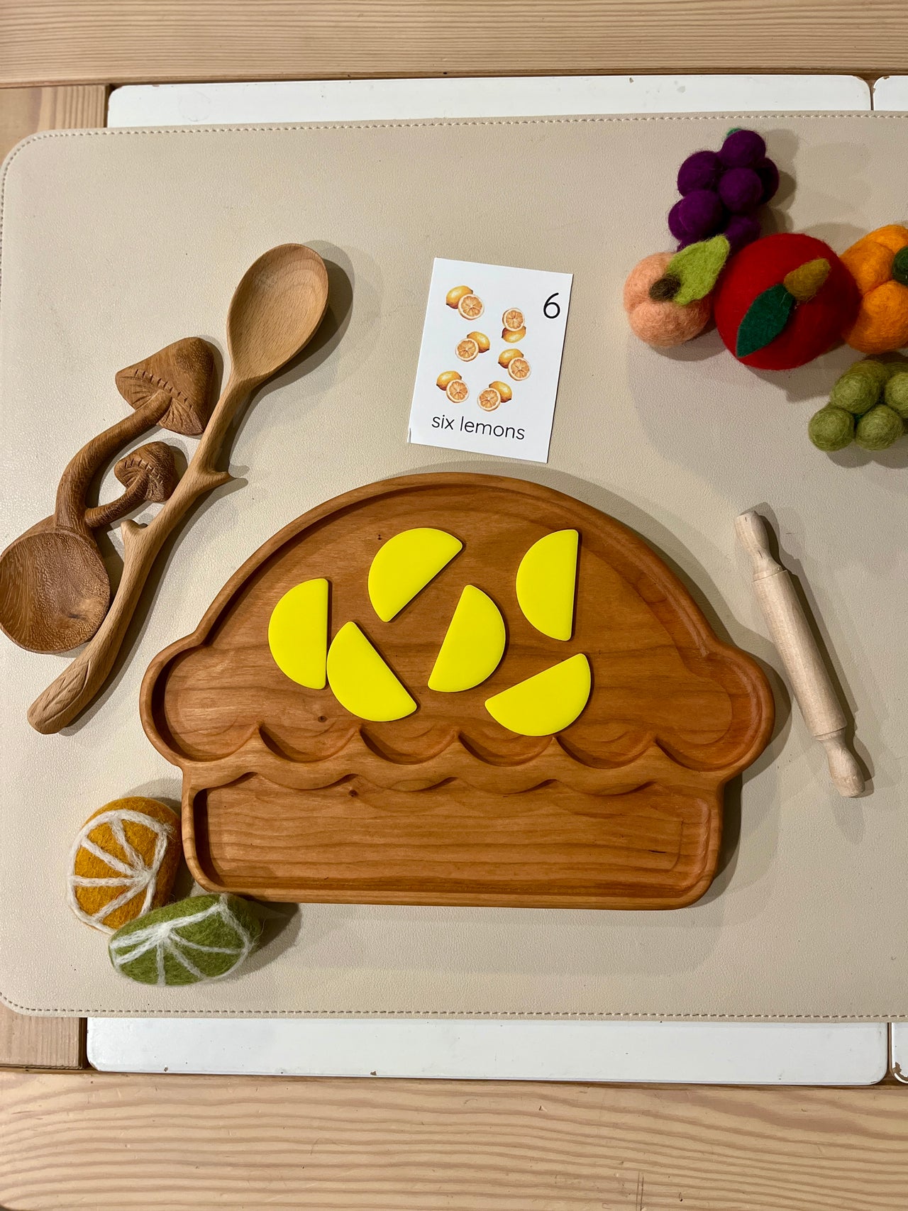 Wooden play food set with a muffin-shaped tray holding six yellow lemon wedges and surrounded by toy cooking utensils and felt fruit.