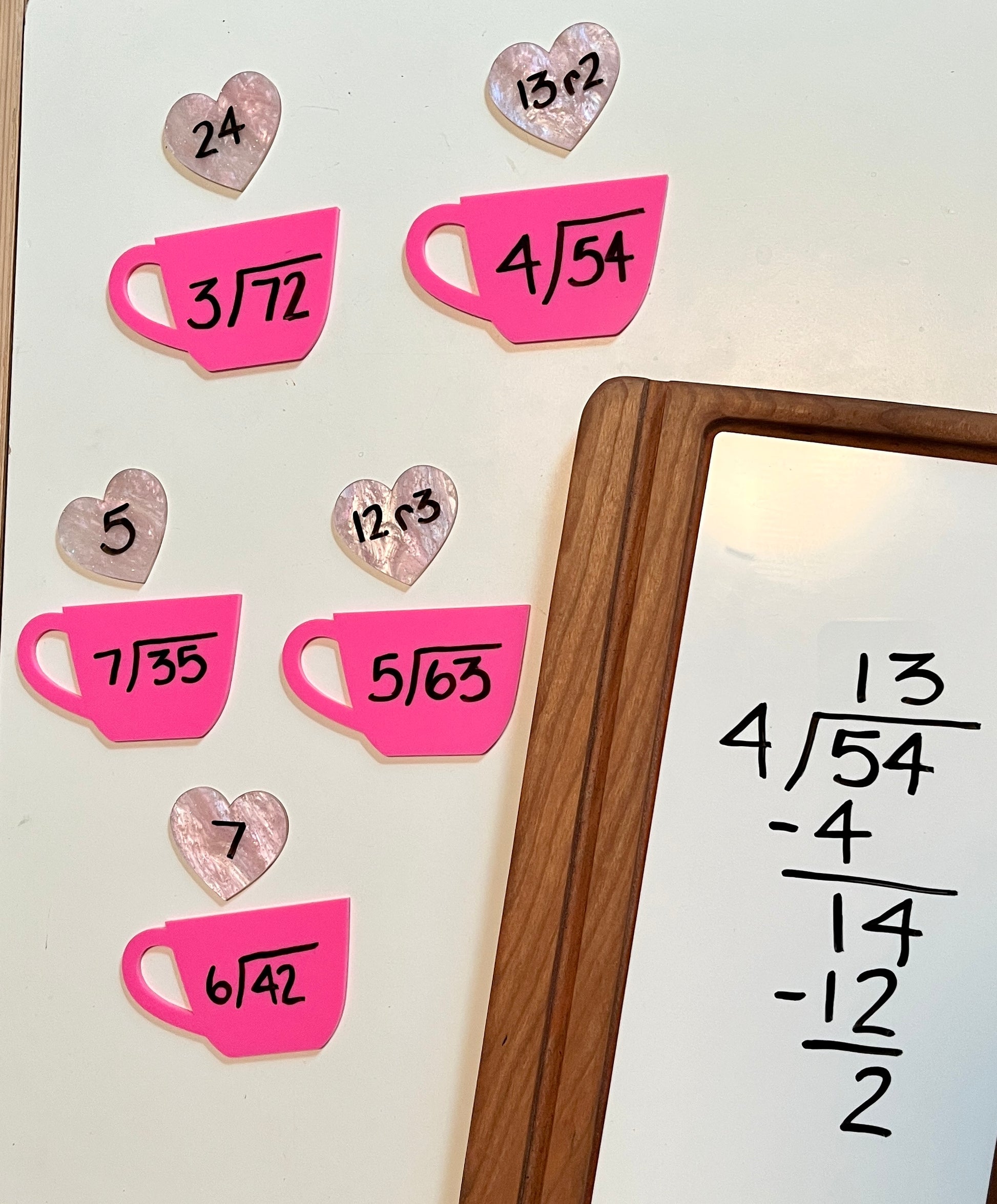 Mathematical equations on pink dry erase shaped like teacups and hearts, with a whiteboard in a wooden tray in the background with the equations on it. 