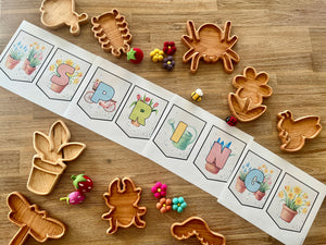 Arrangement of handcrafted wooden Bug Trays in various insect and flower shapes alongside colorful felt pom-poms and a spring-themed educational banner, homeschool resources.