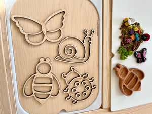 Wooden board with cutouts of butterfly, snail, bee, and ladybug shapes alongside a tray with colorful natural craft materials