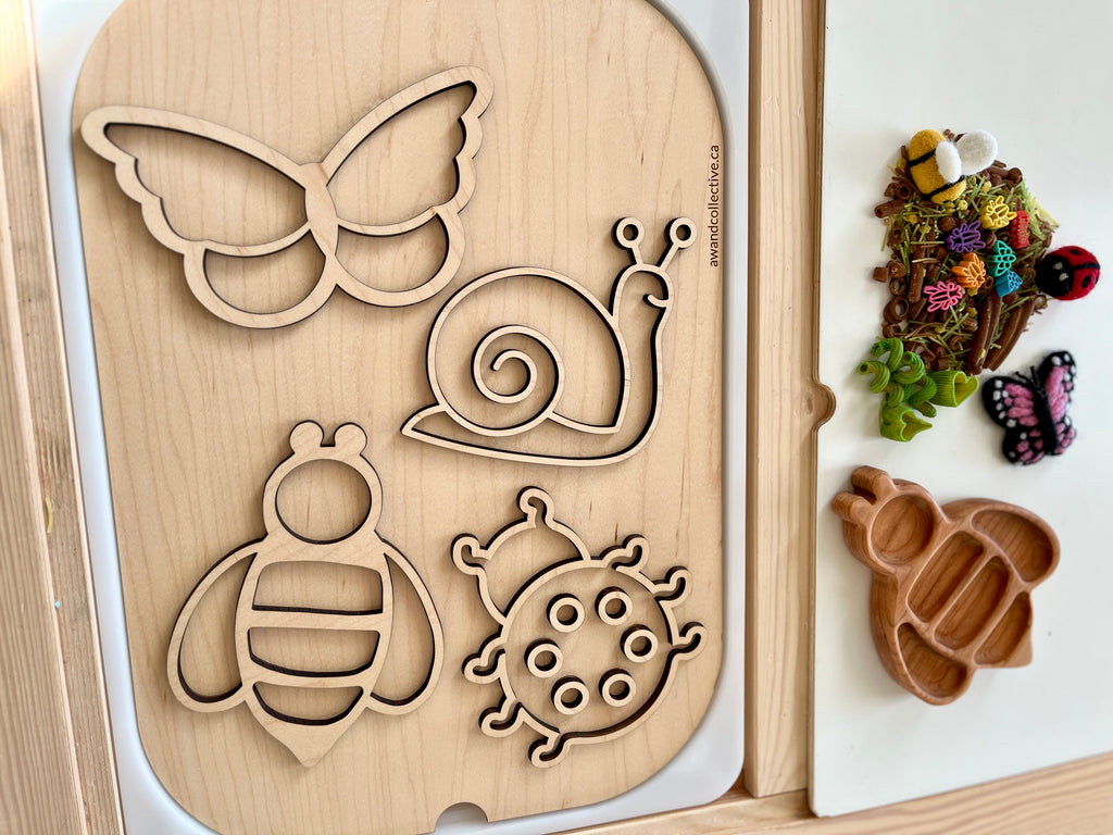 Wooden board with cutouts of butterfly, snail, bee, and ladybug shapes alongside a tray with colorful natural craft materials