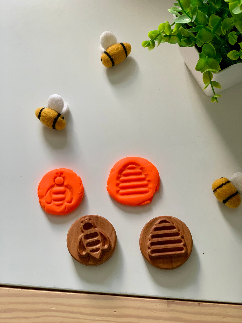 Wooden stamps with bee and hive designs pressed into orange clay, surrounded by three felted bee toys and a green plant on white surface