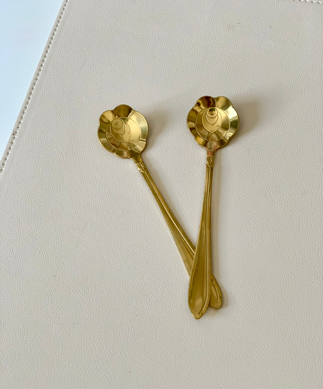 Two gold-toned ornate spoons with scalloped bowl edges resting crossed on a beige textured surface