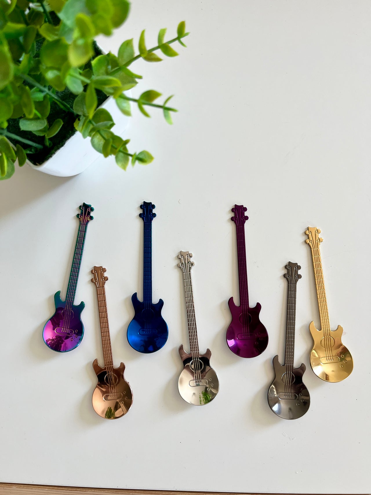 Seven small metallic guitar-shaped objects in various colors arranged on a white surface next to a green potted plant