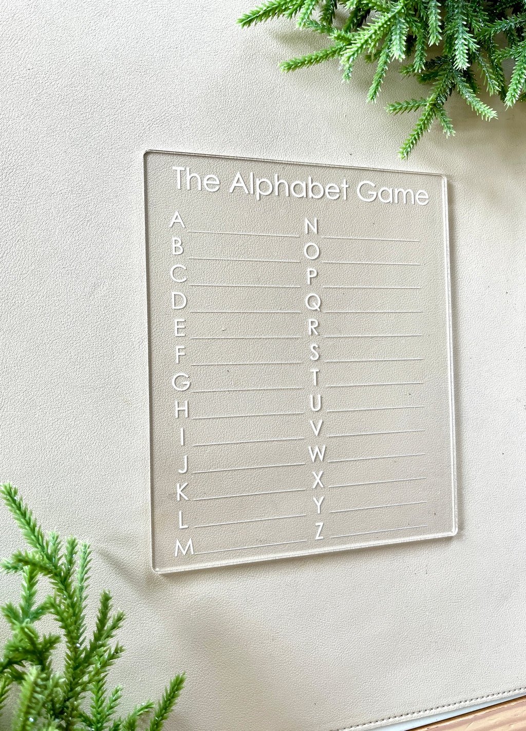 Clear acrylic alphabet game board with engraved letters arranged in two columns on light surface with green foliage accents