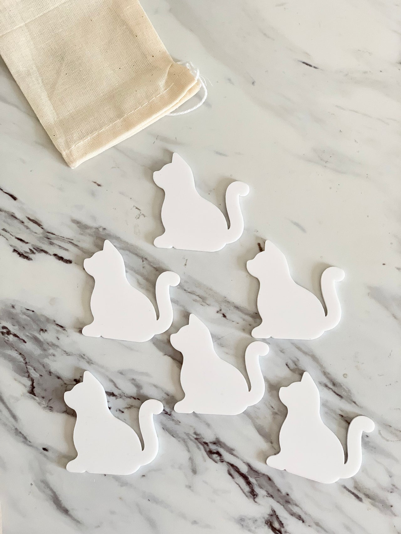 Set of six white dry erase cat-shaped pieces with a small cloth drawstring bag on marble surface