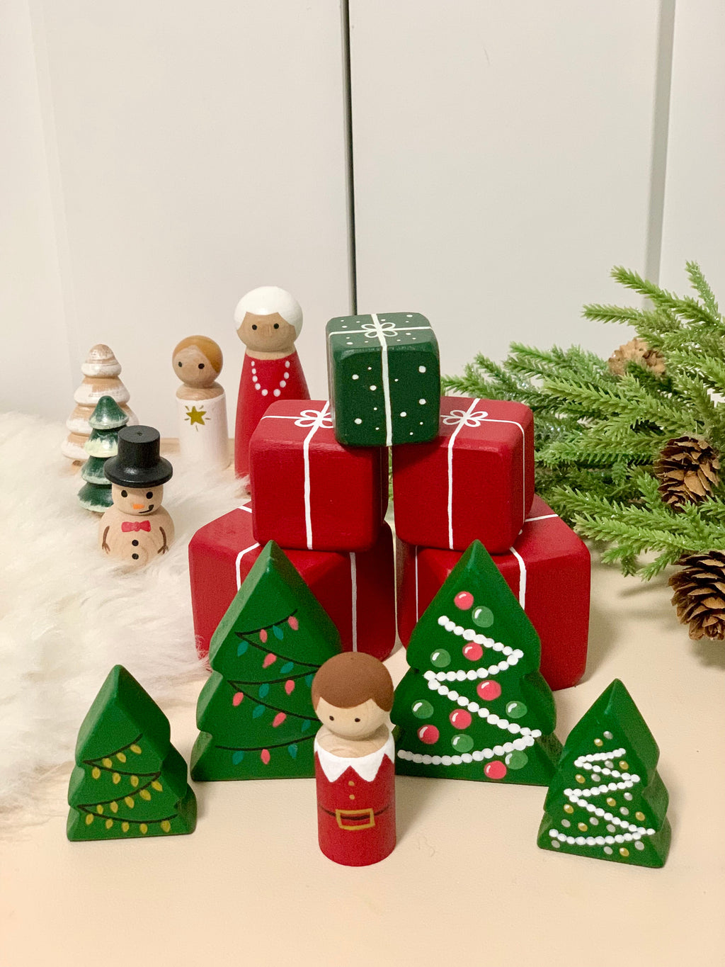 Set of two wooden Christmas tree figurines with painted decorations alongside peg doll and red gift boxes on white surface