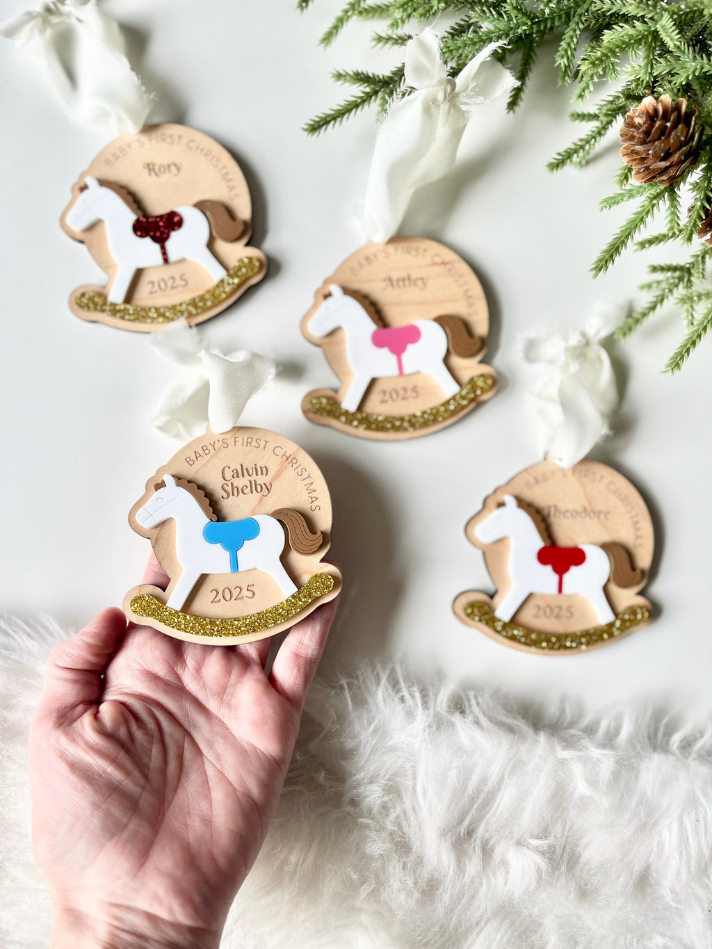 Hand holding wooden rocking horse ornament with colorful saddle and gold glitter base alongside three similar ornaments on white background
