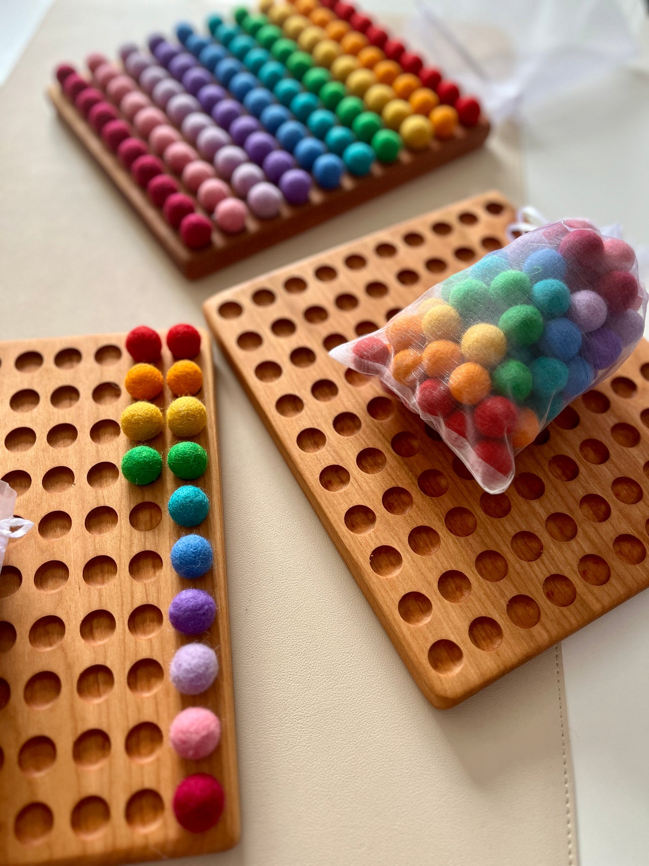 Three wooden hundred sorting boards with smooth engraved holes and colorful felt balls arranged and in a mesh bag on light surface