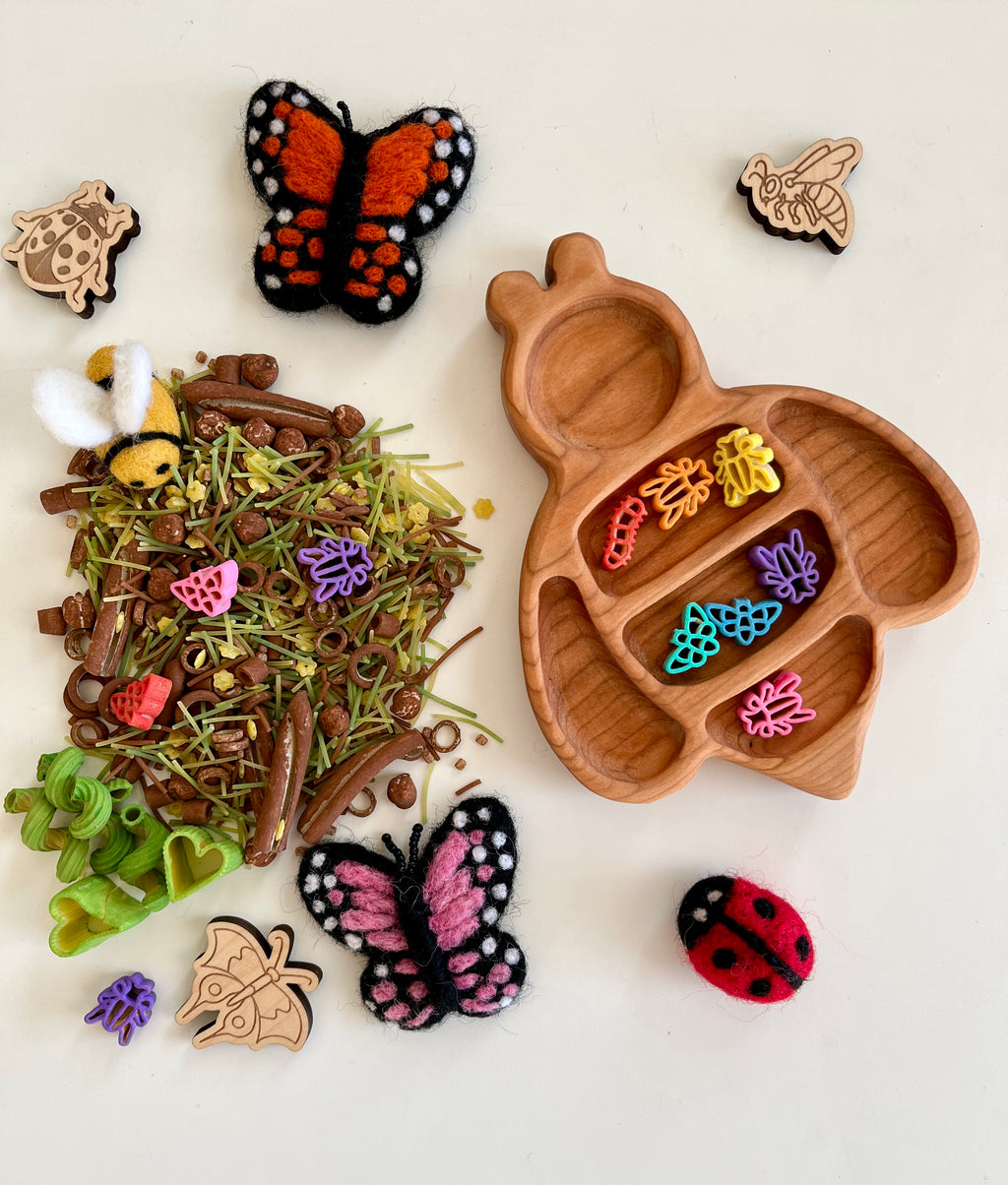 Wooden bee-shaped tray with colorful plastic insect-shaped pieces, felt butterflies, ladybug, and natural craft materials scattered nearby