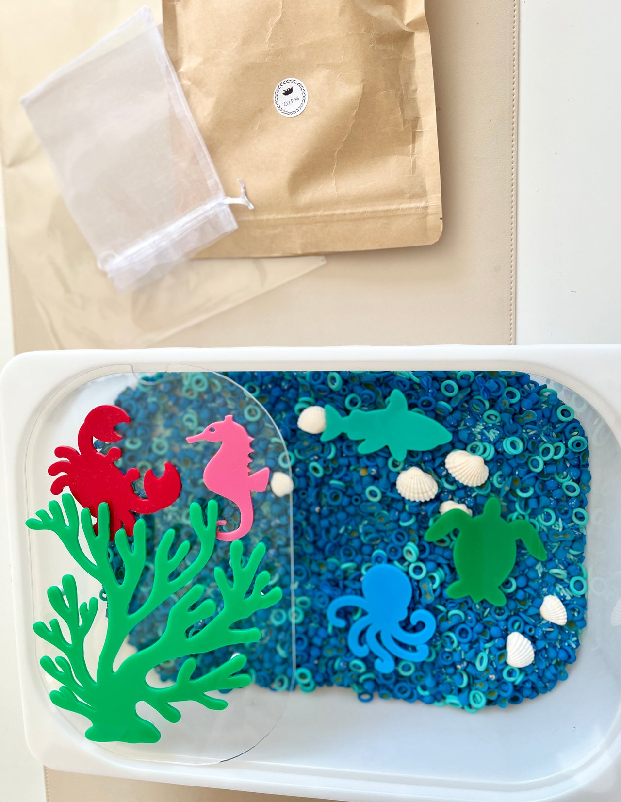 Plastic container with blue circular beads, white seashells, and colorful sea creature shapes on beige surface
