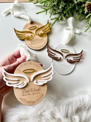 Three angel wing memorial ornaments in gold, white, and bronze tones with personalized names and dates on white surface