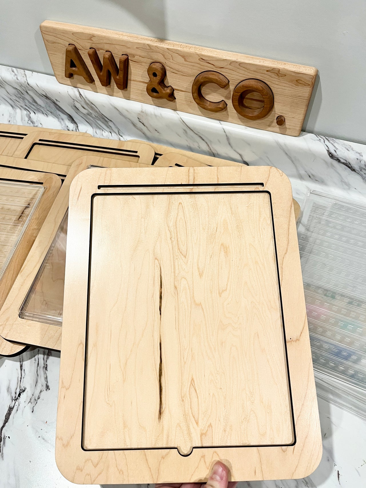 Light wooden tray with a rectangular groove held above other trays on a marble-patterned surface under a wooden AW & CO sign