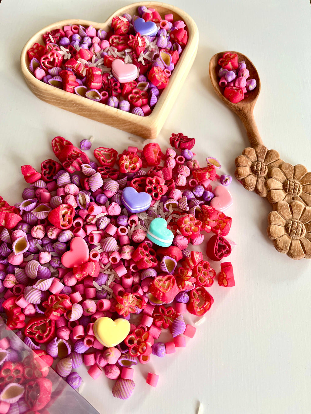 Assorted pink, red, and purple heart and flower-shaped pasta scattered next to a floral-carved wooden spoon and heart-shaped bowl