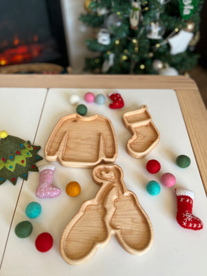 Wooden trays shaped like sweater, mitten, and stocking surrounded by colorful felt balls and small decorative stockings on table