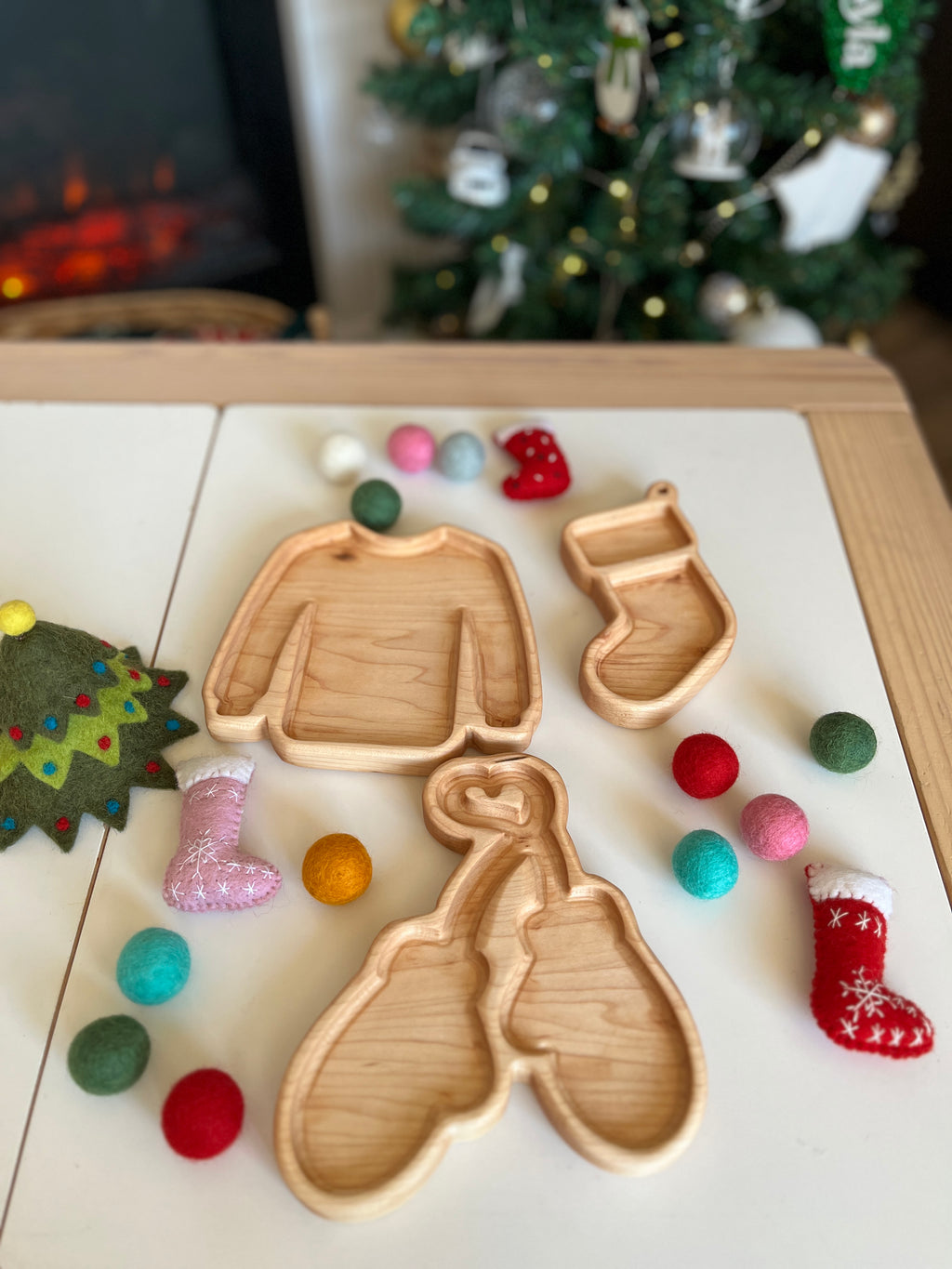 Wooden trays shaped like sweater, mitten, and stocking surrounded by colorful felt balls and small decorative stockings on table