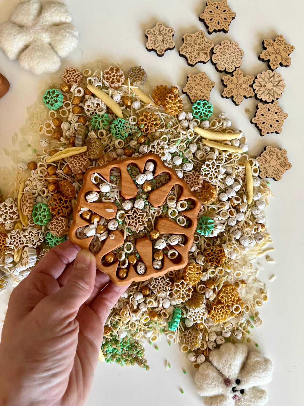 Hand holding wooden snowflake-shaped tray over mixed natural sensory materials with dried botanicals and pasta on white surface