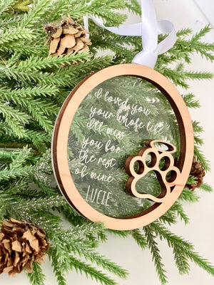 Circular wooden ornament with paw print cutout and sentimental message hanging on pine branches with cones