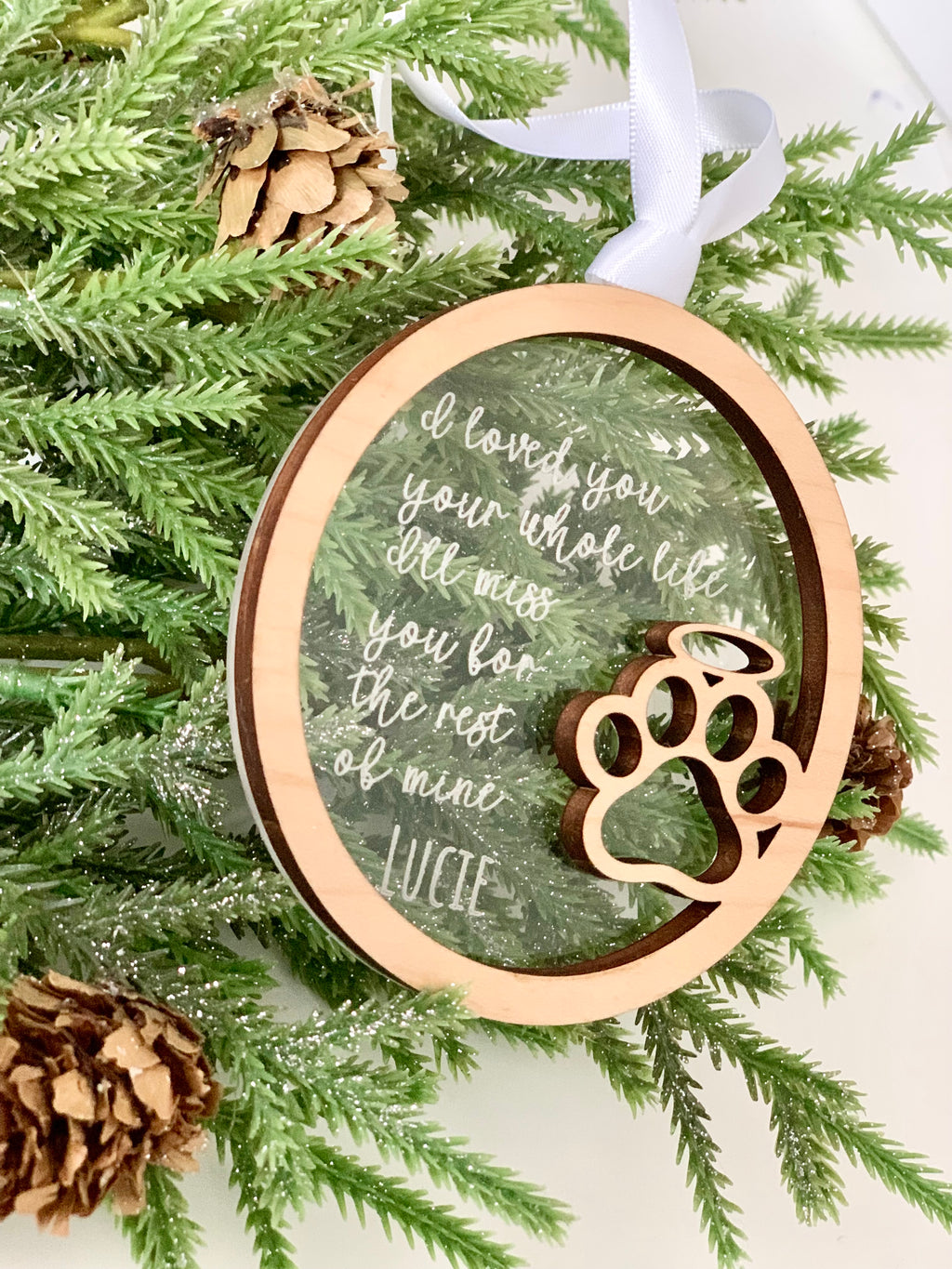 Circular wooden ornament with paw print cutout and sentimental message hanging on pine branches with cones
