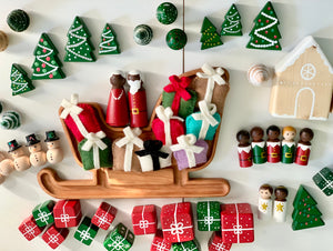 Hand-painted wooden Christmas peg dolls, gift boxes, trees, snowmen, and sleigh on white surface with gingerbread house
