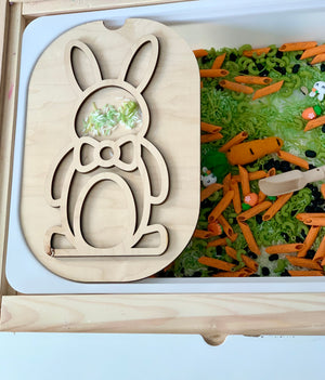 Wooden bunny-shaped stencil resting on a white tray filled with green and orange sensory materials and small toys