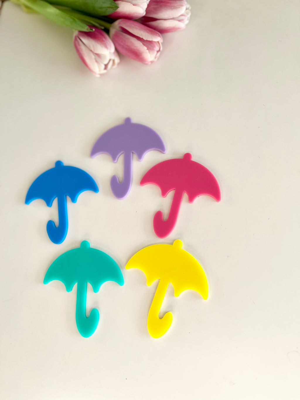 Five colorful umbrella-shaped cutouts in blue, teal, purple, pink, and yellow beside pink tulips on pale surface