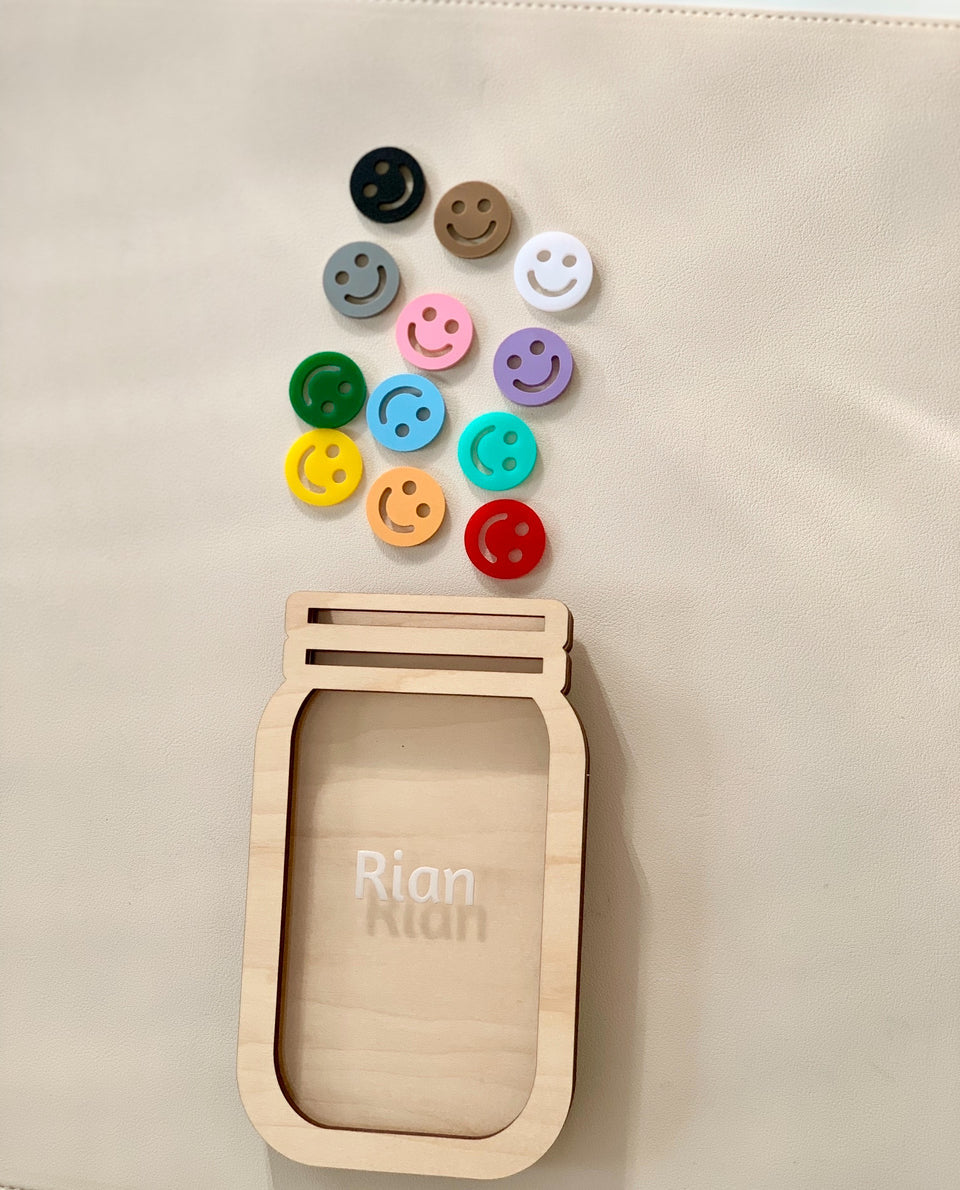 Light wooden jar-shaped frame personalized with the name Rian and colorful smiley face tokens arranged above on a light surface