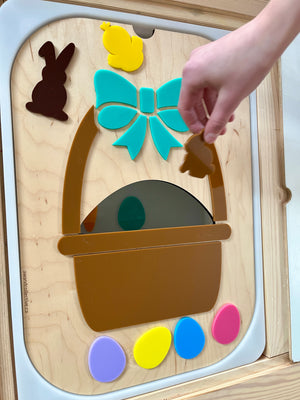 Child’s hand placing brown chick shape into a wooden board with a basket, bow, bunny, chick, and colorful eggs