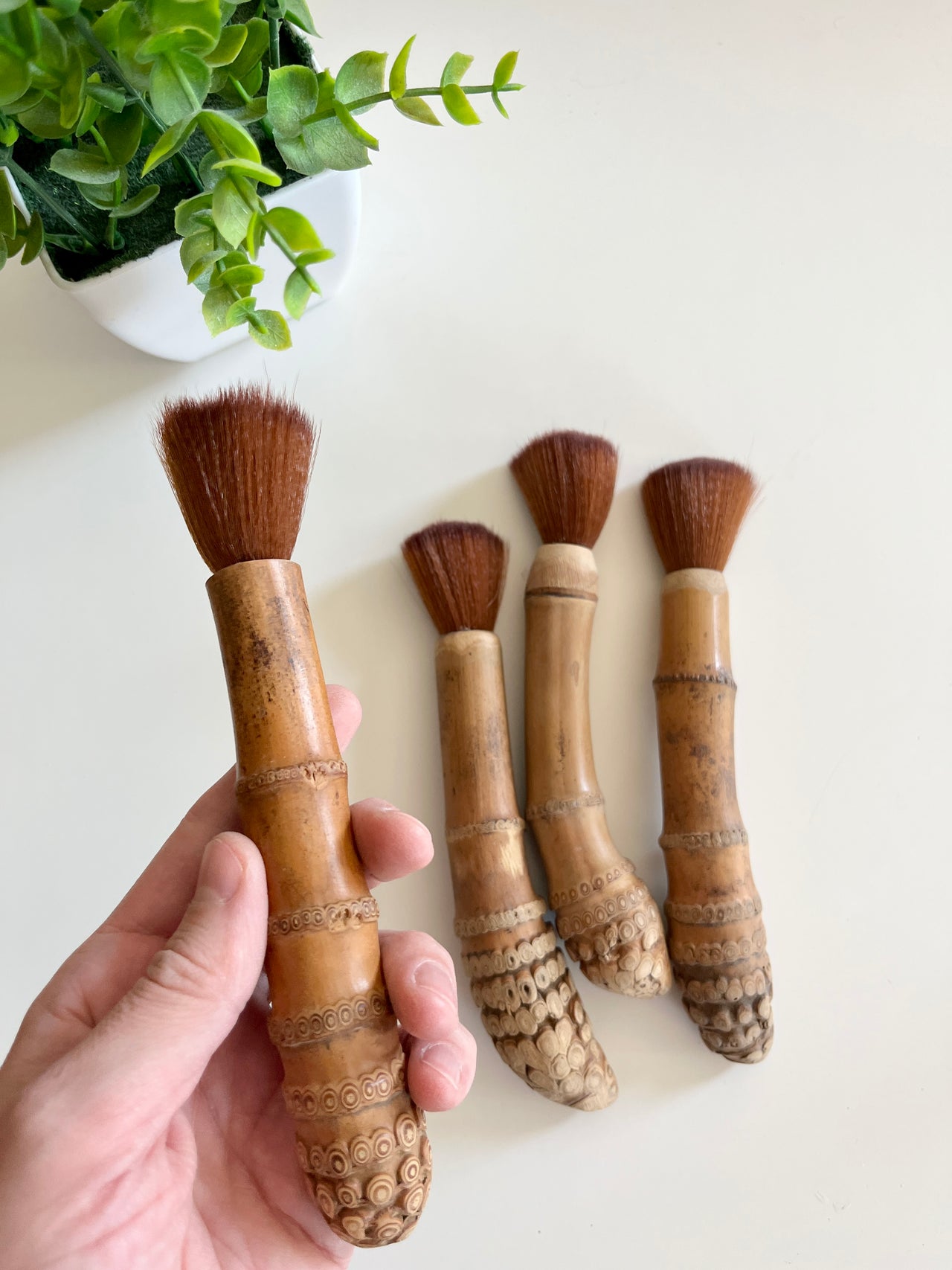 Hand holding a wooden brush with carved handle and natural bristles next to three similar brushes on white surface