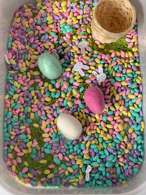 Colorful pastel sensory beans scattered in a container with felt eggs, bunny cutouts, and a small woven basket