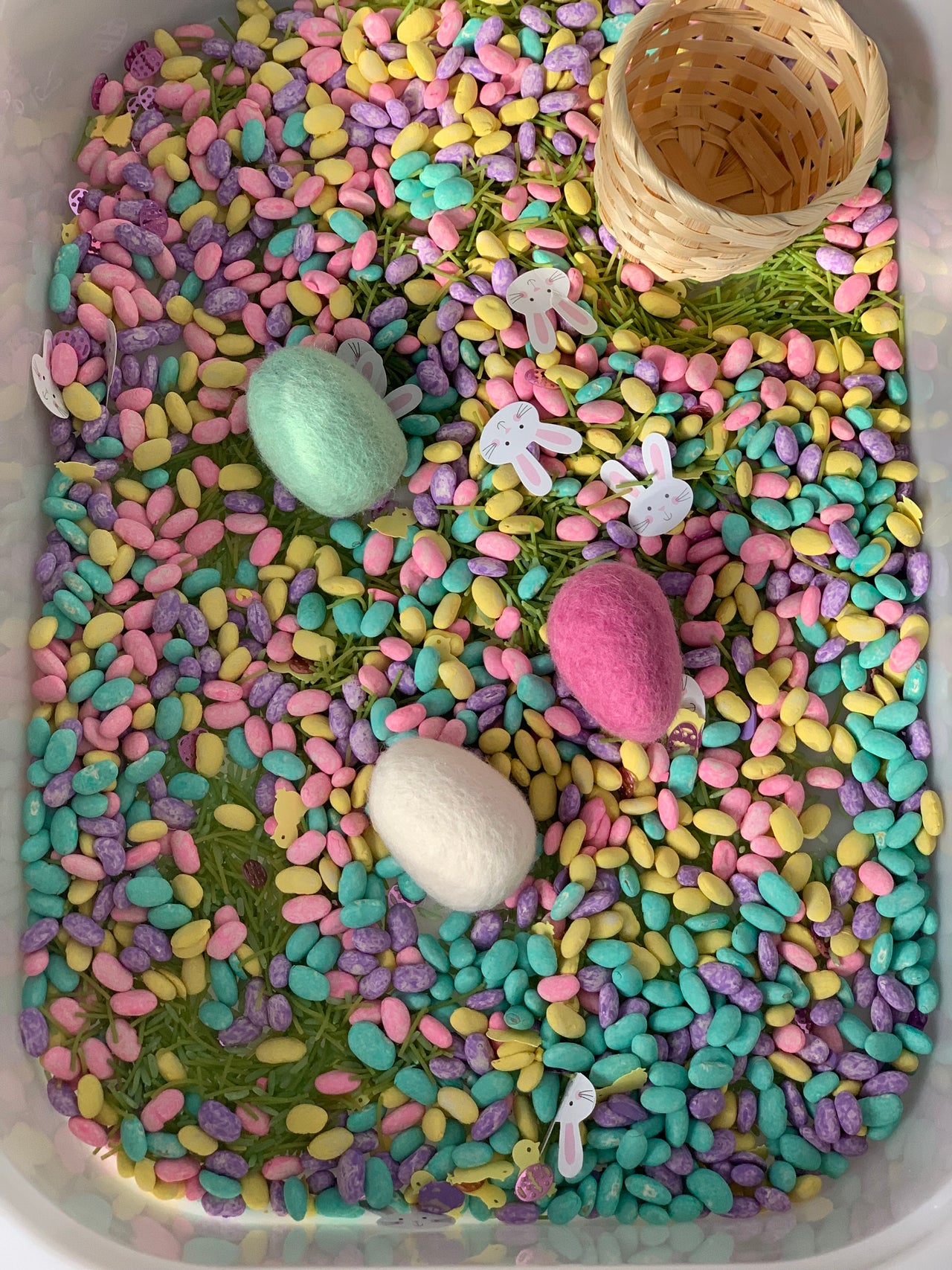 Colorful pastel sensory beans scattered in a container with felt eggs, bunny cutouts, and a small woven basket