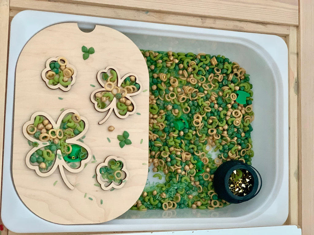 White sensory bin with green and natural pasta, wooden shamrock board, small green shapes, and black pot with gold pieces