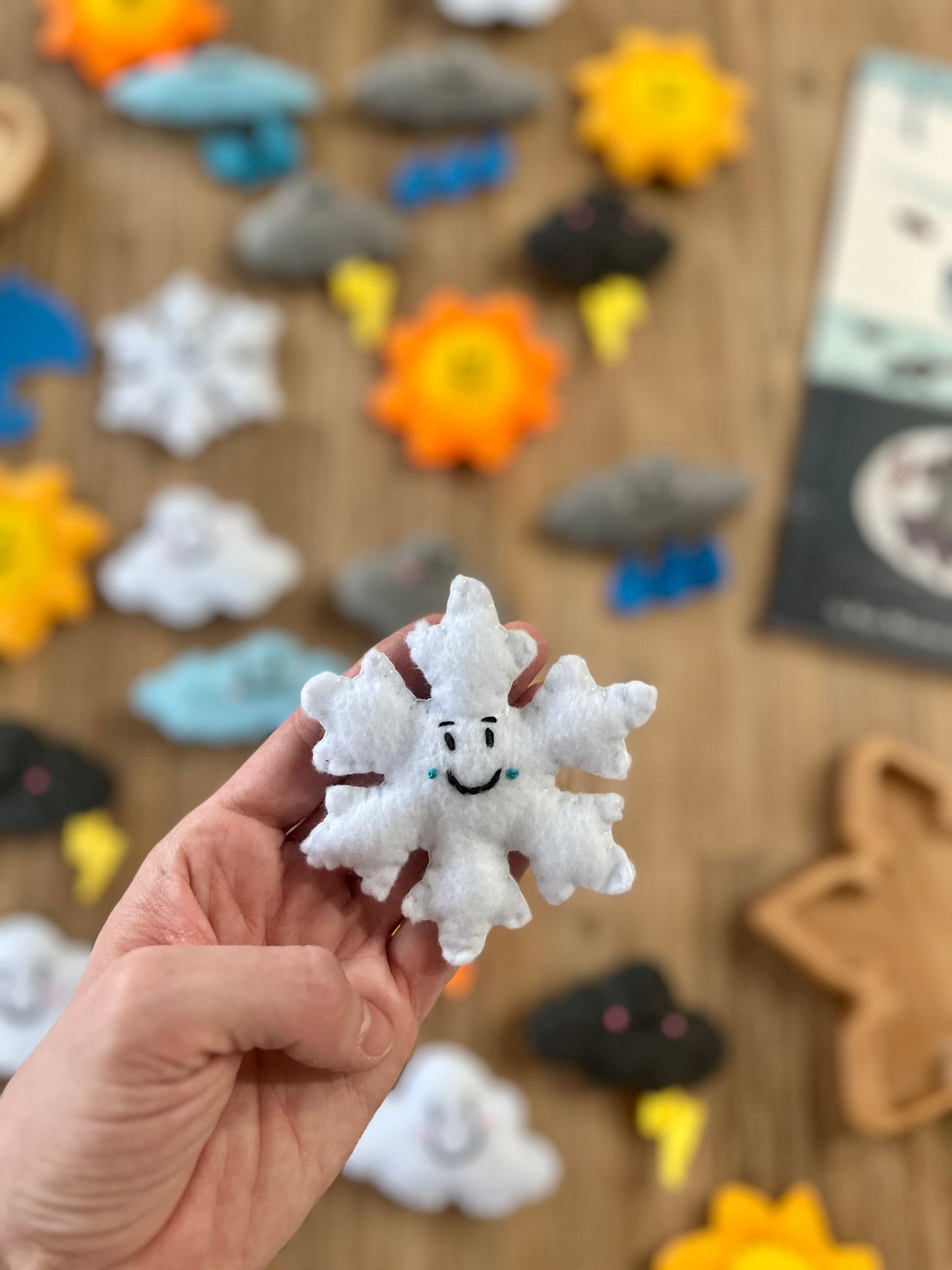 Hand holding a smiling white felt snowflake with blue teardrop details, surrounded by various colorful Felt Weather Shapes, Made in Canada, handcrafted, homeschool, education resources.
