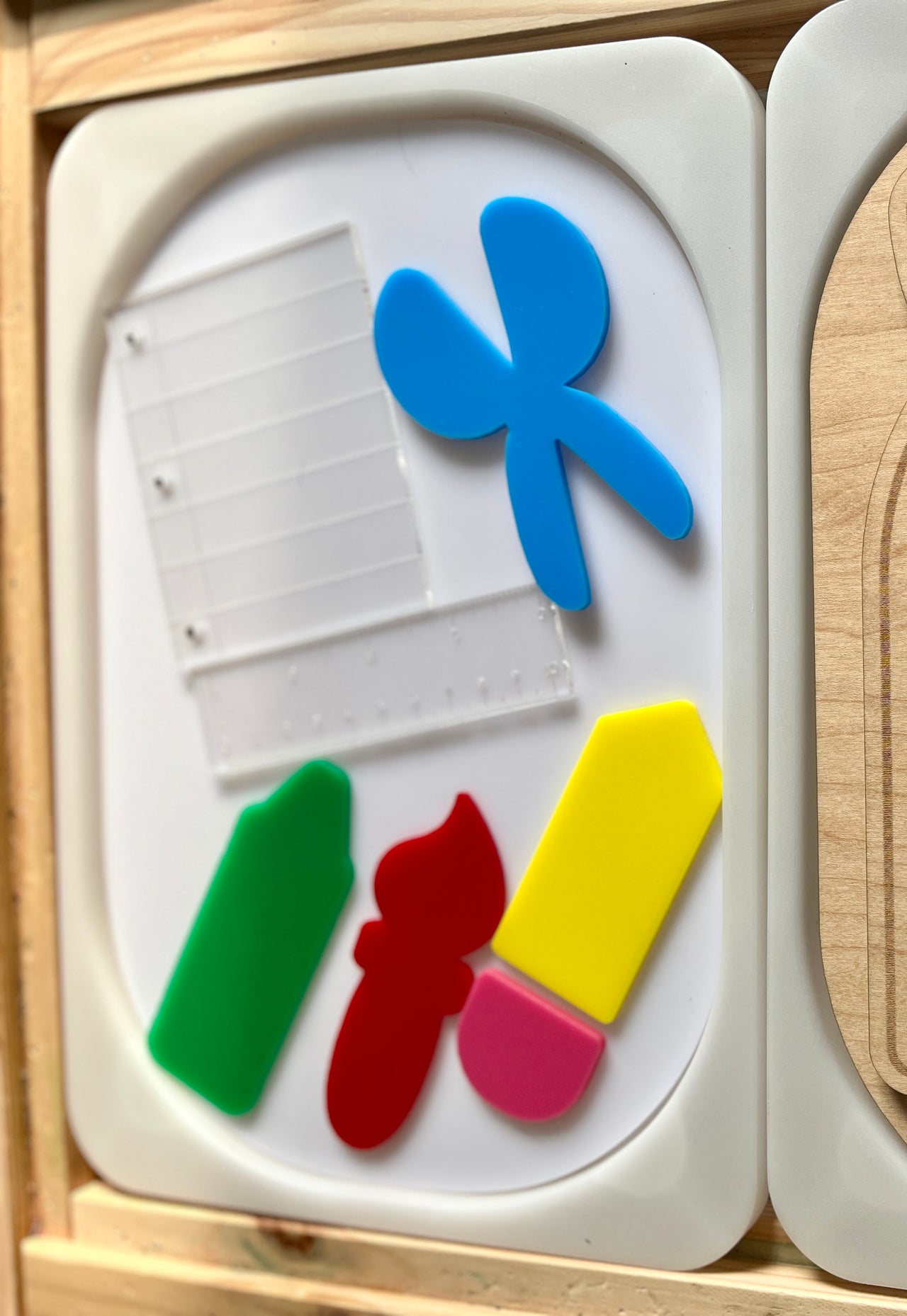 Translucent plastic shapes resembling a fan and measuring tools alongside solid green, red, yellow, and pink geometric pieces on a white tray