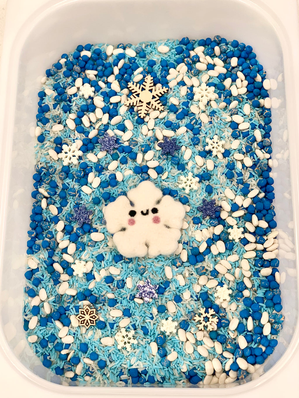Blue and white sensory filler with wooden and felt snowflakes, blue rice, white beans, and a smiling felt snowflake character