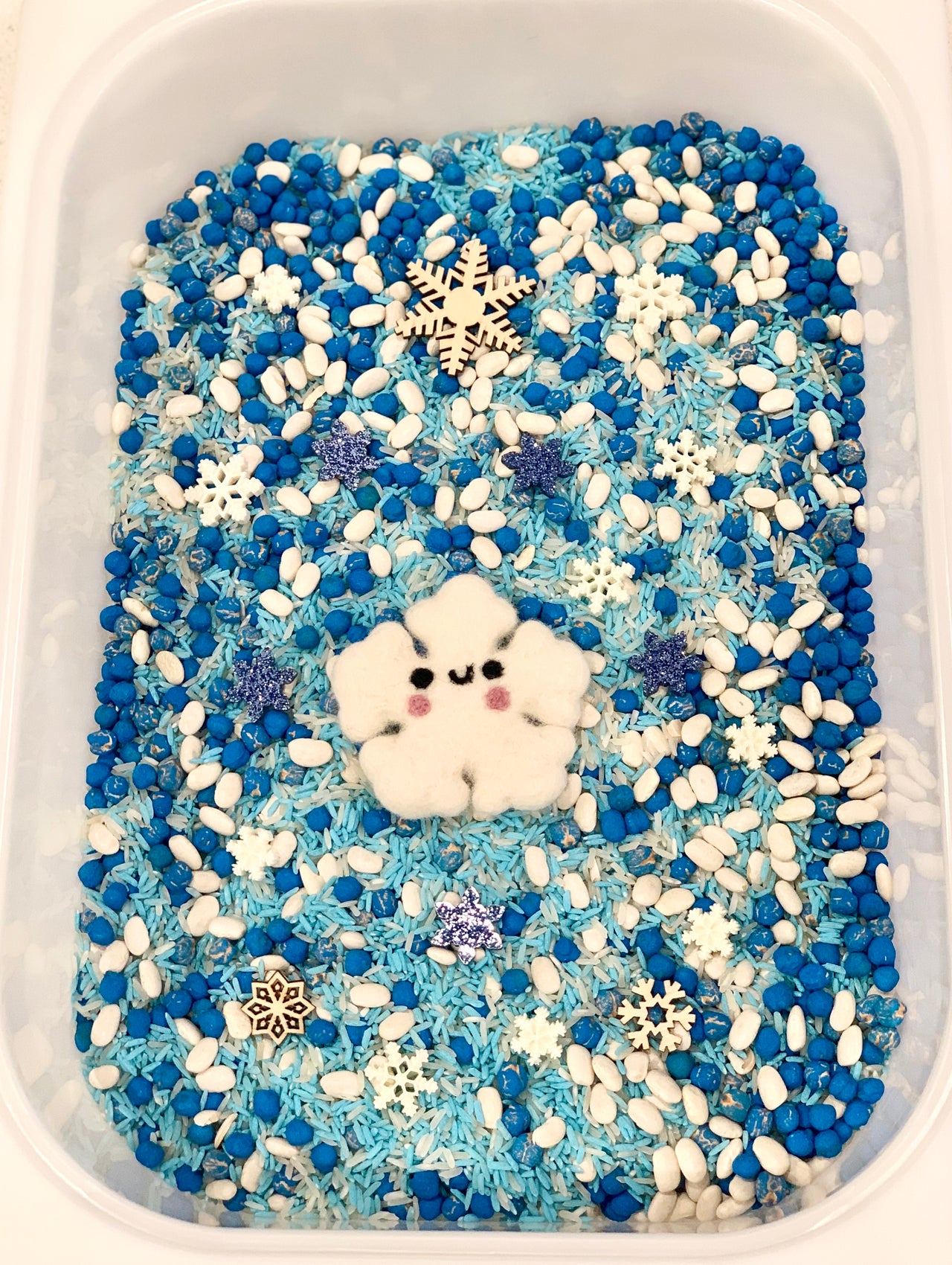 Blue and white sensory filler with wooden and felt snowflakes, blue rice, white beans, and a smiling felt snowflake character