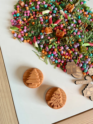 Wooden circular stamps with carved Christmas tree designs near colorful pasta shapes and small wooden holiday cutouts on white table