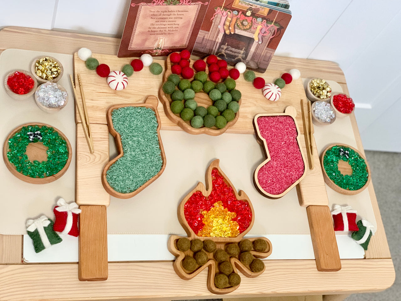 Wooden campfire sensory tray with green and red textured filling, felt garland, and holiday-themed decorations on light wood surface