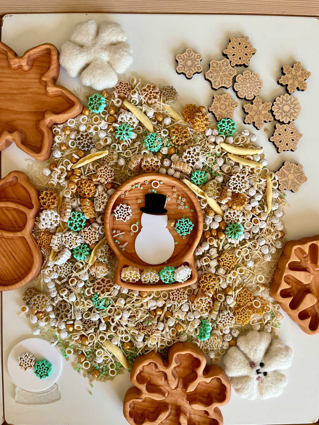 Wooden trays shaped like leaves and snowflakes surround mixed natural sensory materials with snowman and snowflake decorations scattered on table