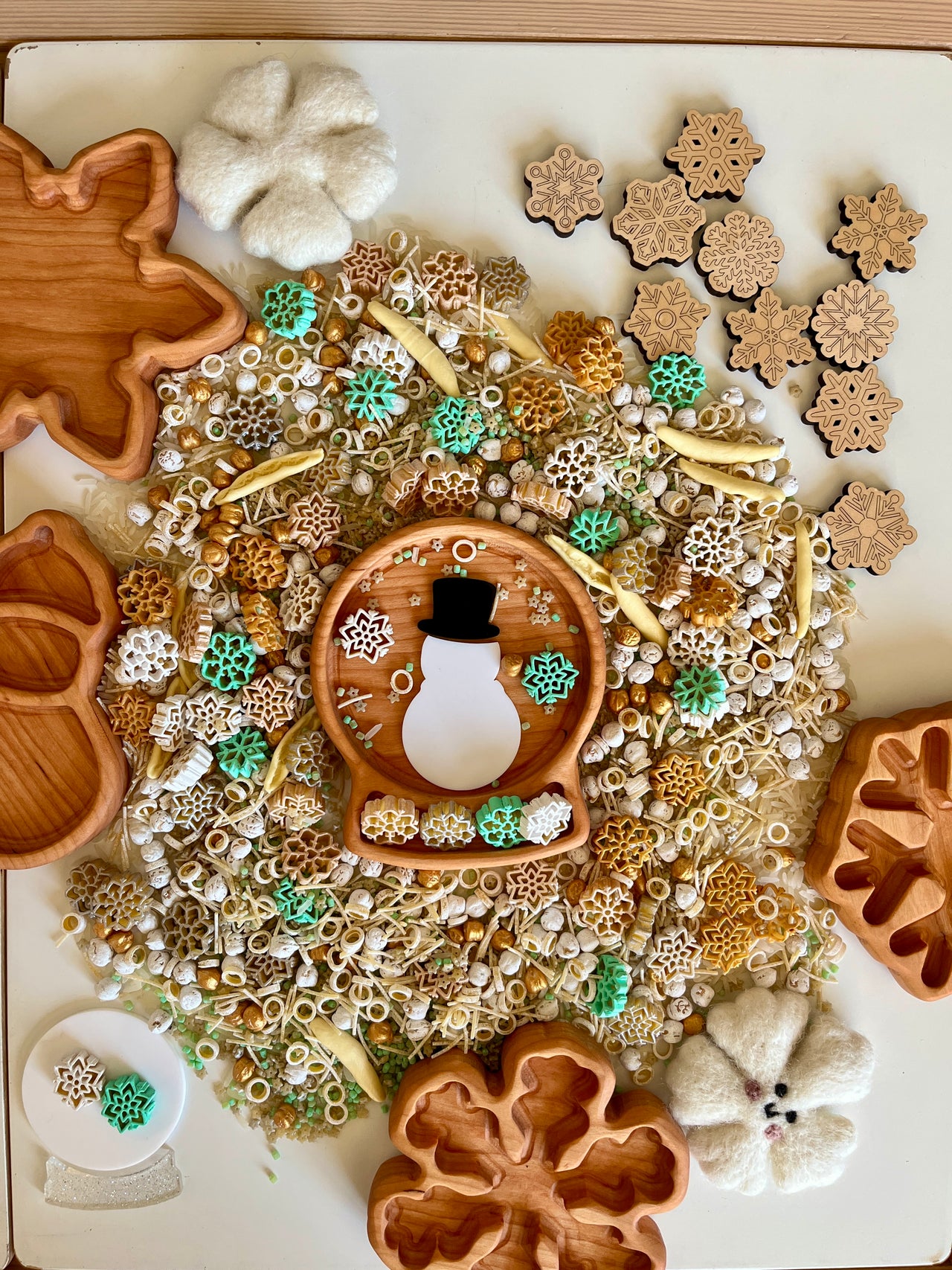 Wooden trays shaped like leaves and snowflakes surround mixed natural sensory materials with snowman and snowflake decorations scattered on table
