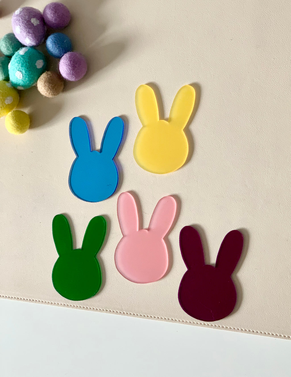 Five colorful bunny-shaped acrylic cutouts in yellow, blue, green, pink, and burgundy on a beige surface with felt balls nearby