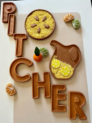 Wooden letter trays spelling "PITCHER" with a wooden pitcher and round tray holding yellow rice, seeds, and felt citrus fruits