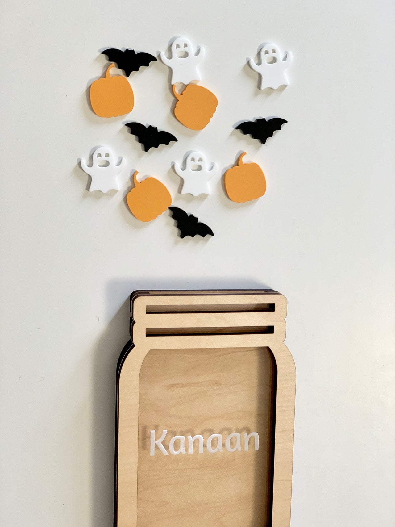 Light wooden jar-shaped frame with the name Kanaan and scattered orange pumpkin, white ghost, and black bat tokens above on a white background