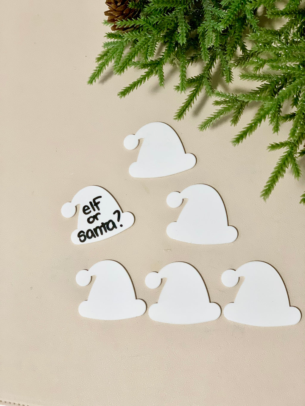 Set of six white dry erase Santa and elf hat shapes with one written "elf or Santa?" on beige surface with greenery above